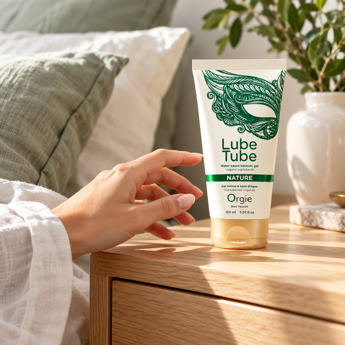 A hand holds a tube of Orgie Lube Tube water-based lubricant gel next to a wooden bedside table in a softly lit bedroom setting.
