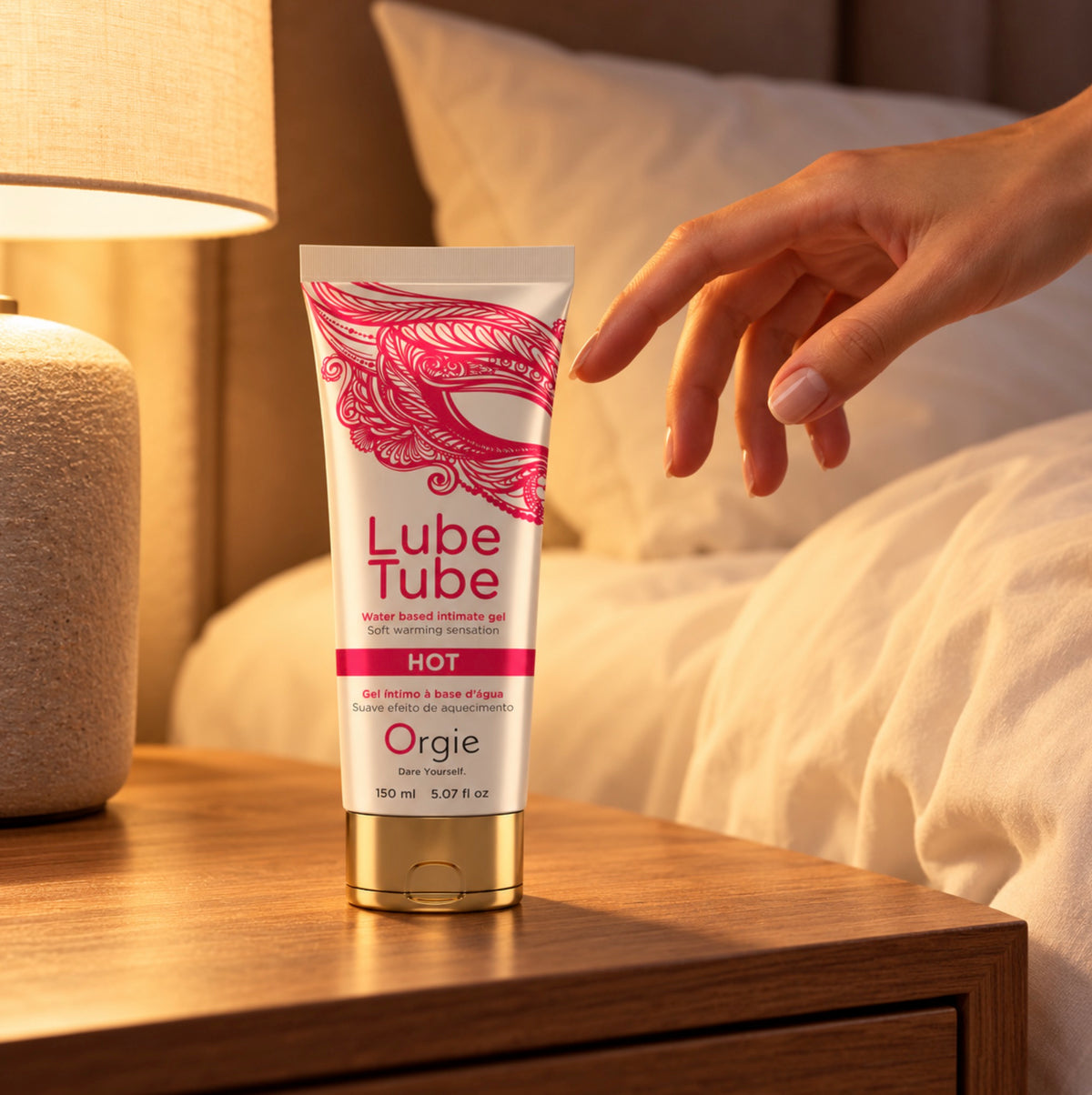 A hand reaches toward a tube of Orgie Lube Tube hot lubricant gel placed on a wooden bedside table next to a lit lamp and bed.